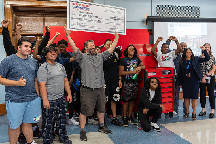 A man in a gray shirt holds an oversized check for $100,000 above his head, surrounded by several high school students and adults.