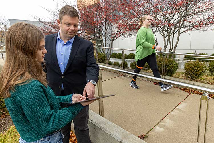 Assistant Professor of Biomedical Engineering Jacob Rammer discusses a mobility test with one student, as another walks by wearing monitoring equipment.