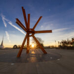 An orange sculpture stands on a concrete plaza with the rays of the sun shining between two of the lower steel beams.