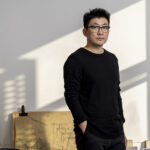 A man wearing a black shirt stands in a room with the slanting outline of a window o the wall behind him.