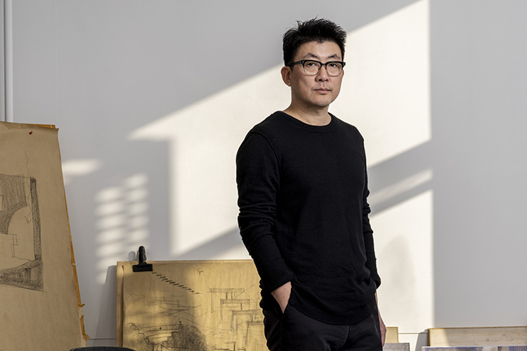 A man wearing a black shirt stands in a room with the slanting outline of a window o the wall behind him.