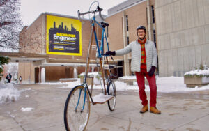 A man stands next to his very tall bicycle on the plaza between two buildings