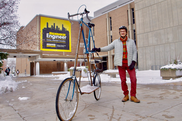 A man stands next to his very tall bicycle on the plaza between two buildings