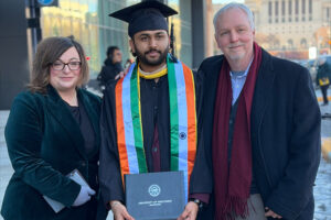 A man and a woman stand on each side of a man wearing graduation regalia. All are standing outside on a sidewalk.