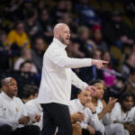 A man wearing a white shirt points while standing on the sidelines of a basketball court.
