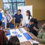 Four college students stand while talking with several adults sitting at a table that has printouts of webpages.