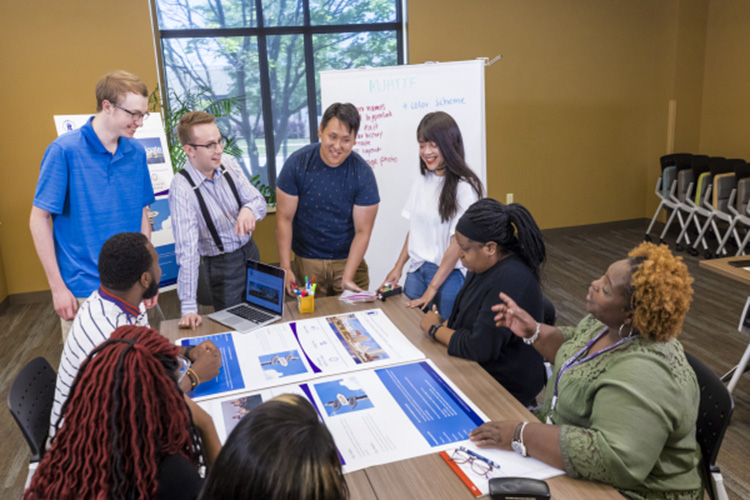 Four college students stand while talking with several adults sitting at a table that has printouts of webpages.