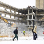 Several people walk while bundled in winter clothes past a building being deconstructed. Two large machines tear at the walls of the building, which is mostly torn down.