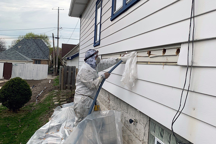A technician in a white protective suit fills holes in an exterior wall of a home with insulation from a hose.