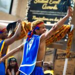 Two dancers wearing colorful clothing raise their hands in the air and wave a stick with ribbons.