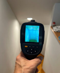 A hand is pointing a thermal imaging tool down a hallway and the display depicts areas of the wall where the temperature varies.