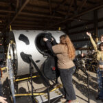 A woman pours food scraps from a black bucket into a large composting drum, while another woman throws flower petals in the air in celebration.