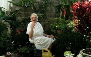An older woman sits in a greenhouse, looking toward the camera.