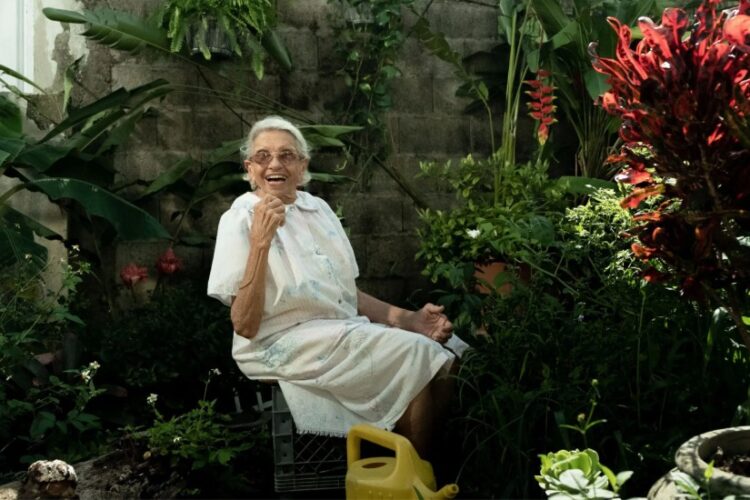 An older woman sits in a greenhouse, looking toward the camera.