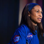 A close shot shows the head and shoulders of a woman wearing a blue uniform with NASA patches on it