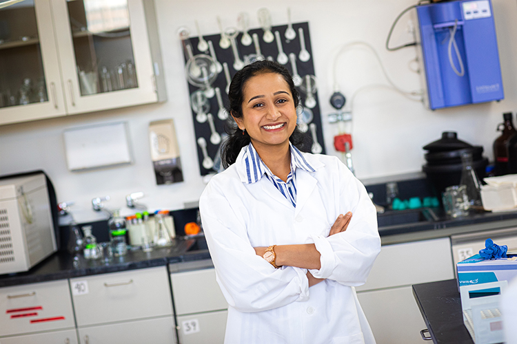 A woman in a white lab coat stands in a lab with crossed arms.