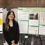 A woman in a black outfit smiles at the camera while standing in front of her research poster board.