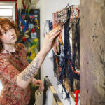 A woman uses a brush to apply paint to a canvas, which also has swirls of fabric attached.