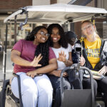 Three people sit in the back seat of a golf cart, smiling and waving to the camera.