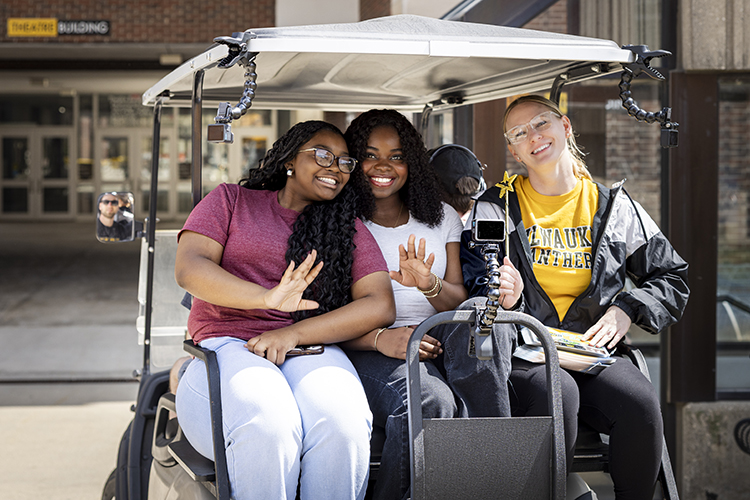 Three people sit in the back seat of a golf cart, smiling and waving to the camera.