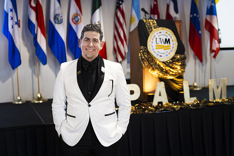 A man wearing a black shirt and white sport coat stands in front of a line of flags and a UW-Milwaukee logo.