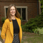 A woman wearing a black shirt and yellow blazer stands outside in front of a red brick building.