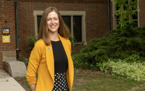 A woman wearing a black shirt and yellow blazer stands outside in front of a red brick building.