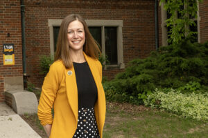A woman wearing a black shirt and yellow blazer stands outside in front of a red brick building.