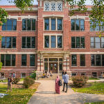 A red brick university building stands in front of several college students amid grass and trees.