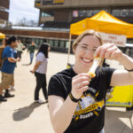A woman holds a cheese curd up to the camera and pulls it apart, stretching the melted cheese. She is standing outside on a sunny day.