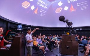 A few dozen people sit in a planetarium, where images of stars and s'mores are projected on the domed ceiling.