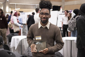 A man in a brown sweater holds a glass plaque in his hands that reads "2025 Panther Edge Student Employment Award."