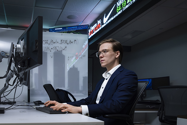 Jack Linge sits at a computer terminal, with a stock ticket board above his head