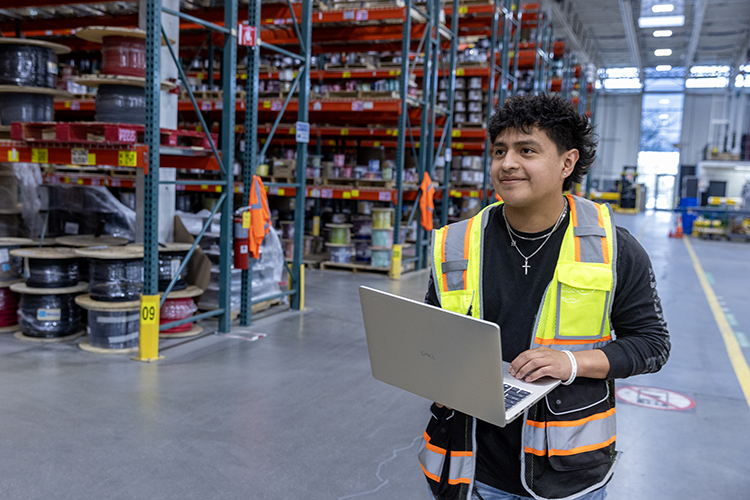 Mason Middaugh stands in a warehouse wearing a safety vest and holding a clipboard
