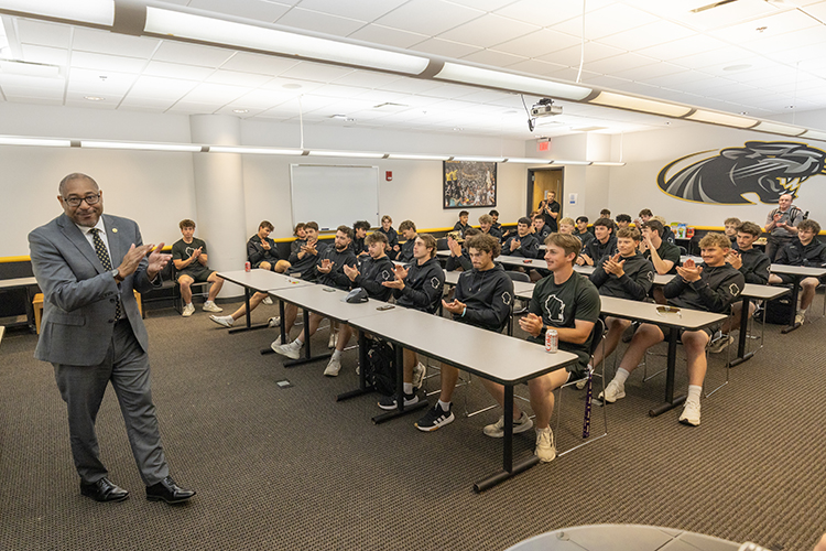 A man stands at the front of a conference room while baseball players fill the seats behind desk. All are clapping.