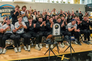 A couple dozen men sit on folding chairs on a gym floor. All are clapping and cheering