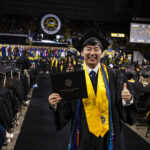 A man in commencement regalia gives a thumbs up and shows his new diploma to the camera while walking down an aisle of graduates.