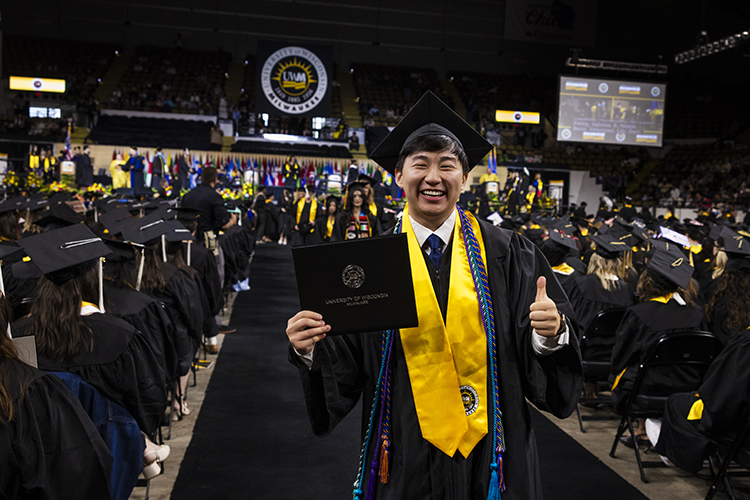 A man in commencement regalia gives a thumbs up and shows his new diploma to the camera while walking down an aisle of graduates.