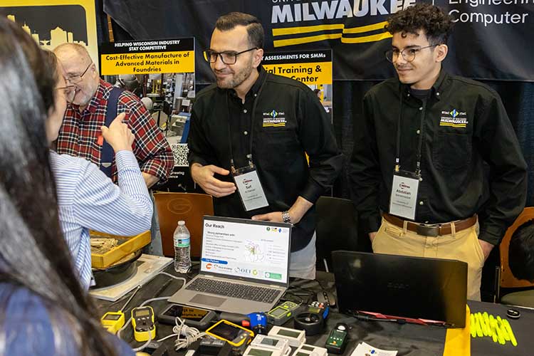Two men wearing black shirts talk with visitors to an exhibition booth.