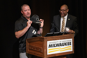 A man holds a baseball cap with a logo of the state of Wisconsin, which he prepares to give to another man behind a podium.
