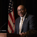A man wearing a suit and tie speaks at a podium, with an American flag in the background.