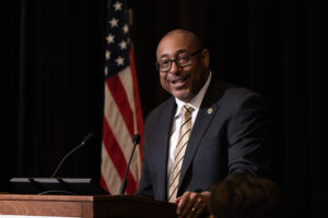 A man wearing a suit and tie speaks at a podium, with an American flag in the background.