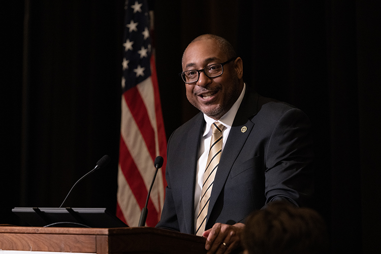 A man wearing a suit and tie speaks at a podium, with an American flag in the background.