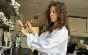 A woman in a white lab coat works with liquid in a beaker in a lab.