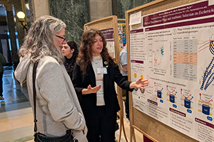 A woman in a suit coat gestures toward a poster board while explaining her research to another woman.