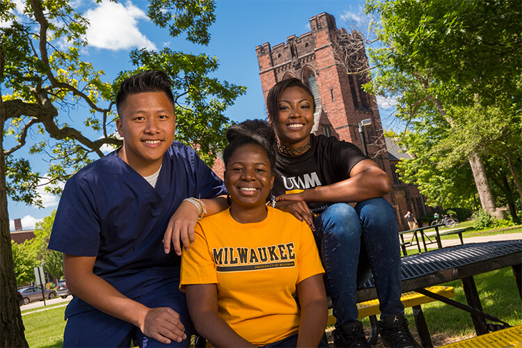 Three students sitting on campus in front of Chapman Hall. One in nursing uniform and two in UWM apparel