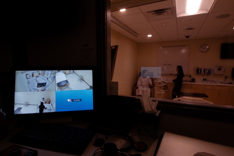 watching the patient and speech student through the simulation glass with the computer in the foreground showing cameras of the ICU