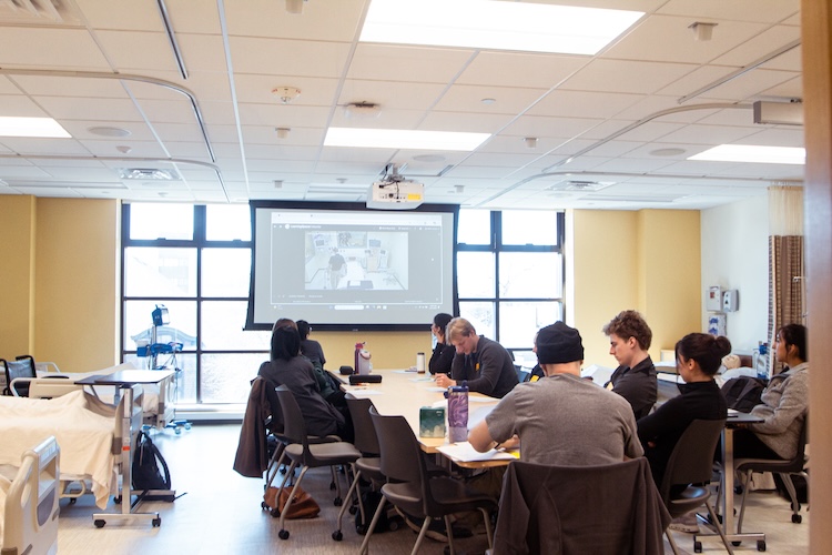 group of student sit working at a table while watching the simulation take place on a giant screen in the room