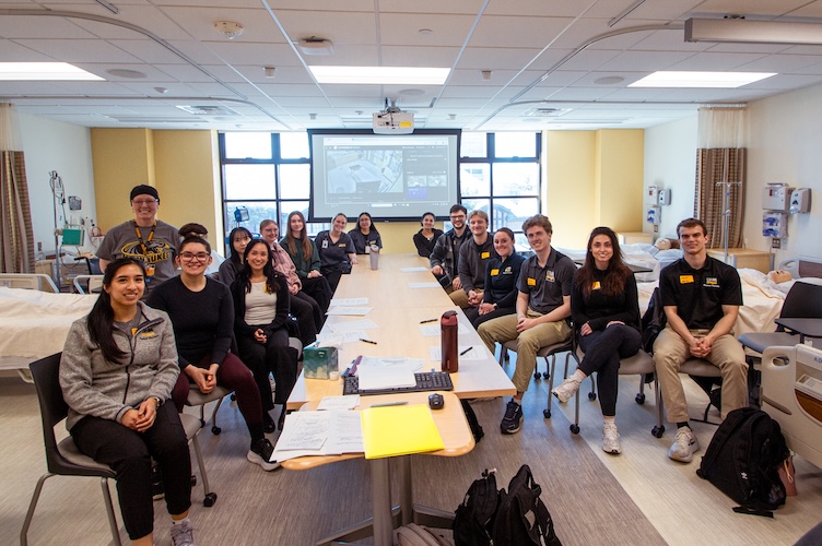 group of nursing, pt, speech students, and professor sit in the skills lab smiling for the camera