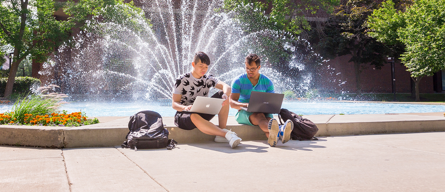 Two students sitting in front of the UWM Fountain using laptops